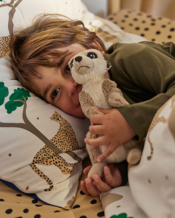 Un niño está abrazando una suricata de peluche en una cama con un edredón y una funda de almohada SANDLÖPARE en blanco y azul oscuro.