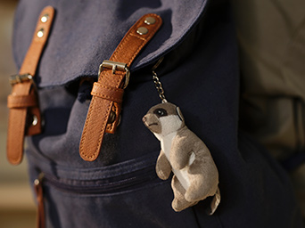Una suricata mini beige de la colección SANDLÖPARE cuelga como mascota en una mochila azul con correas marrones.