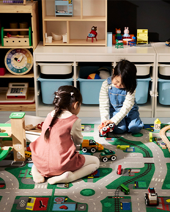 Dos niños jugando con coches de juguete sobre una alfombra de carreteras en un cuarto de juegos organizado con contenedores y estantes