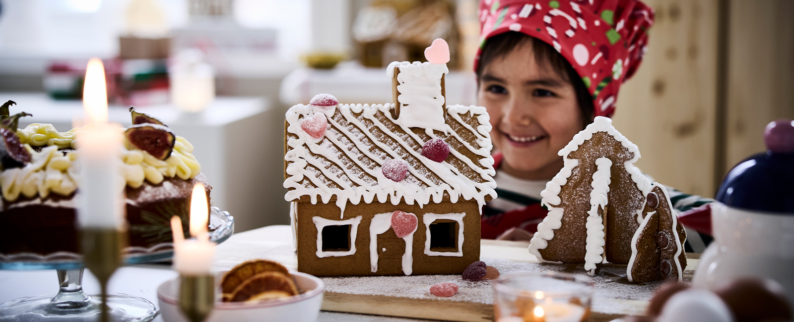 Child decorating gingerbread house with white icing and colorful candies in festive kitchen setting with candles and holiday treats