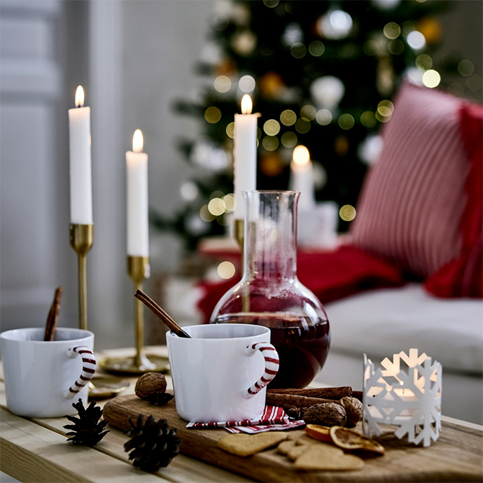 White candles in brass holders, hot drinks in striped mugs, and holiday decorations arranged on wooden surface with Christmas tree in background