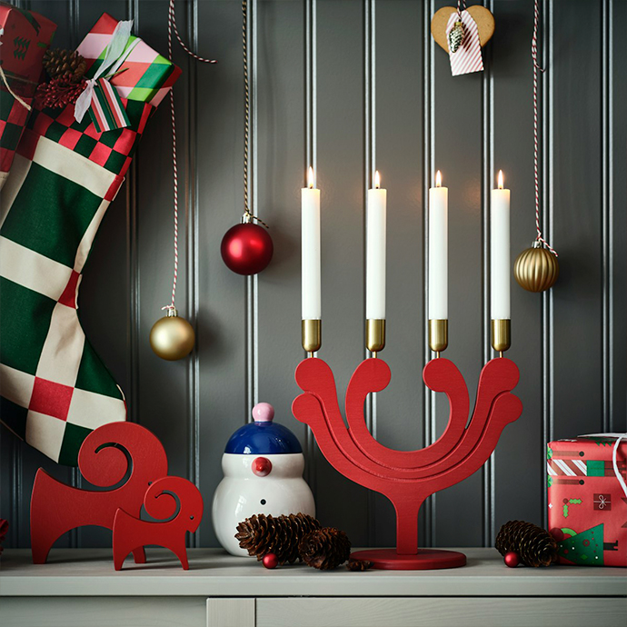 Decoraciones navideñas sobre estante blanco contra pared de paneles oscuros. Candelabro rojo de madera con cuatro velas blancas, adornos navideños colgando de cuerda negra, calcetín a rayas y cajas de regalo
