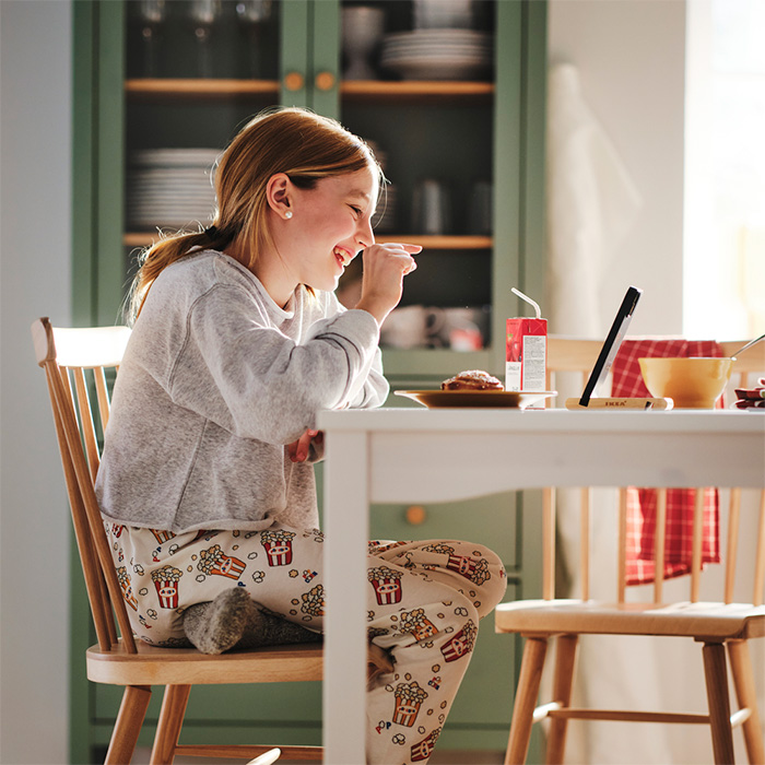a young girl in pyjamas, sitting at the table eating breakfast and looking at her tablet at the same time