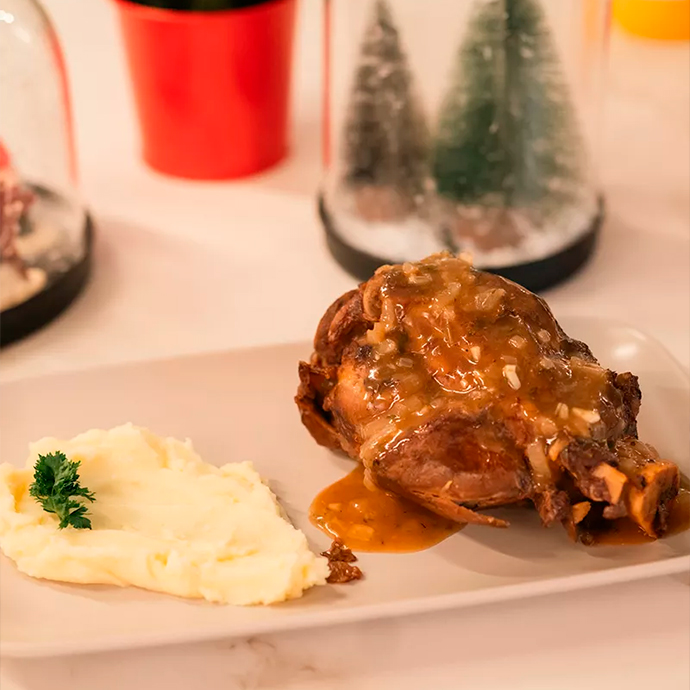 Roasted pork knuckle with brown sauce served on a rectangular white plate, accompanied by mashed potatoes.