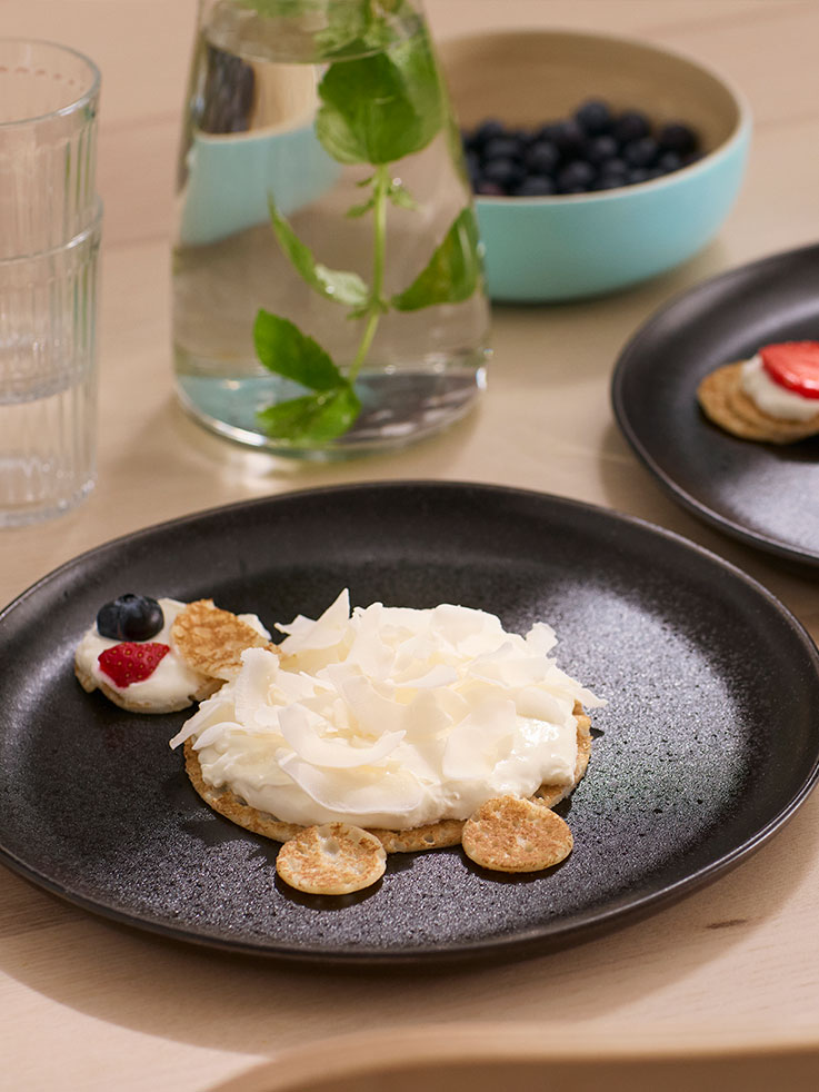 A black plate with pancakes, whipped cream and fruit, cut in smaller pieces and laid out to look like a sheep.