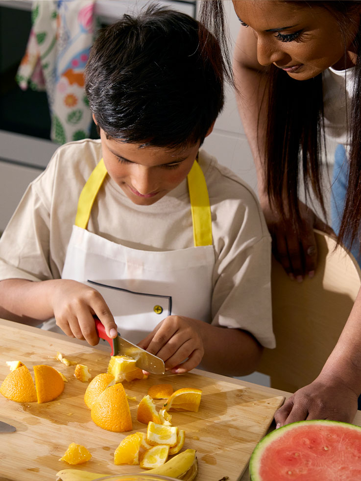A little boy carefully cutting an orange with the help of SMÅBIT knife, the mother watching from the side.