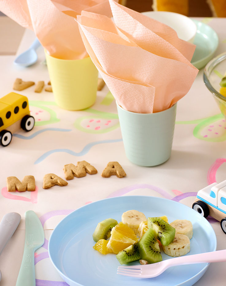 A table with pastel coloured KALAS plates and cutlery, plastic cups with napkins and “mama” spelled out with letter biscuits.