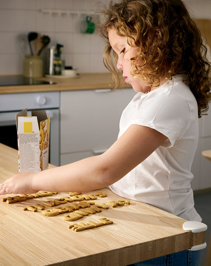 A little girl using KAFFEREP biscuits to spell different names on the kitchen counter. UNDVIKA corner bumper in place.
