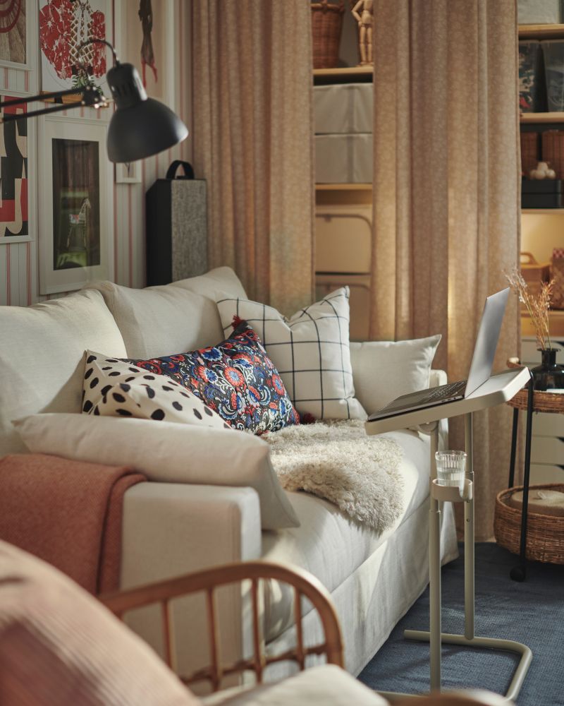 A HYLTARP sofa-bed with some extra cushions on it, including a flowery SKOGSKORN cushion, stands in a living room.