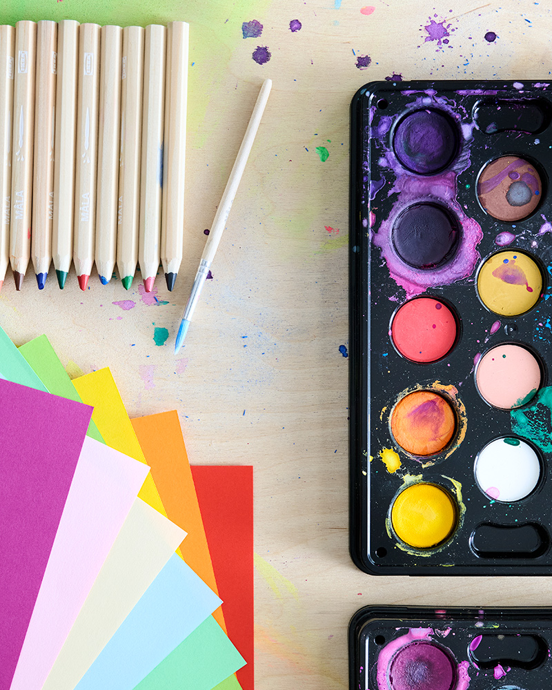 Coloured paper, a set of MÅLA pencils and watercolour box, all laying on a wooden table. Picture shot from above.