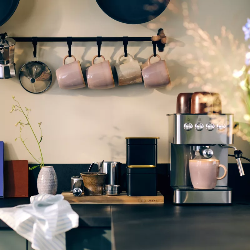 Rustic countertop with coffee maker, FÄRGKLAR mug, and mugs hanging on a wall rail.