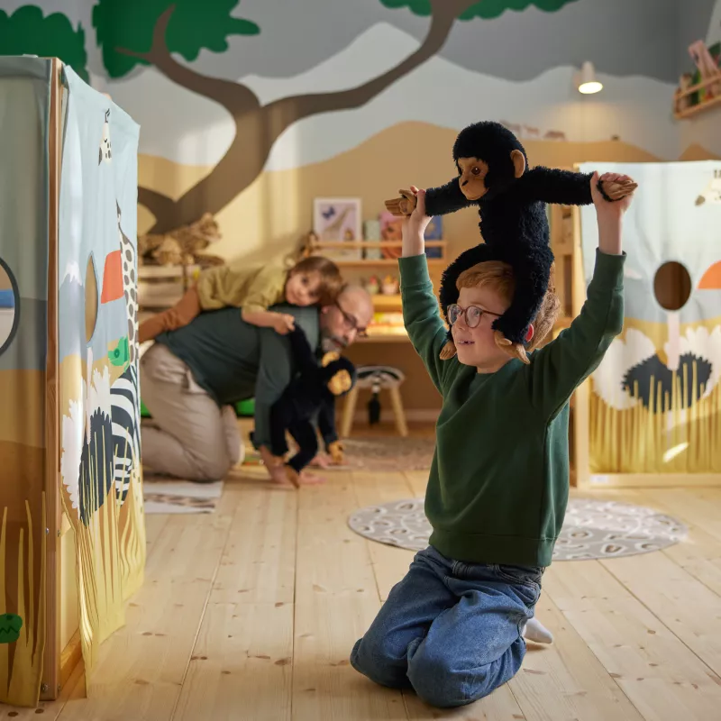 A child sits on their knees in a savannah themed room while carrying a SANDLÖPARE soft toy chimpanzee on their shoulders.