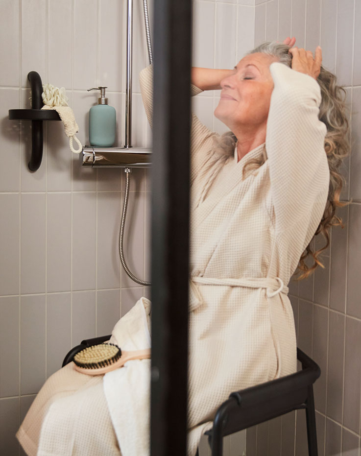  Through the glass shower door: a person in a robe sits  on a grey BÄSINGEN shower chair in anthracite, against  tiled wall.