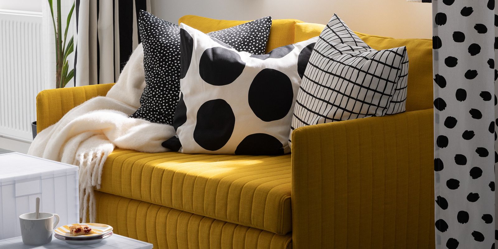 A throw and three cushions, including one with a BLÅMUNKAR cushion cover with large black dots, lie on a FRIDHULT sofa-bed.