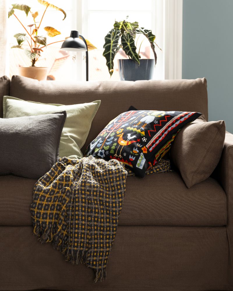 A FLIKBRUNÖRT throw and some cushions, including one with a multicolour TRATTKAKTUS cushion cover, lie on a HYLTARP sofa.