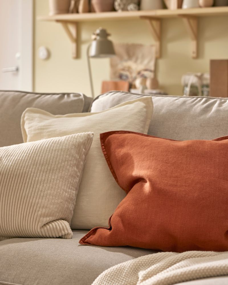 A throw and three cushions, including one with a red brown DYTÅG cushion cover, lie on a light beige EKTORP sofa.
