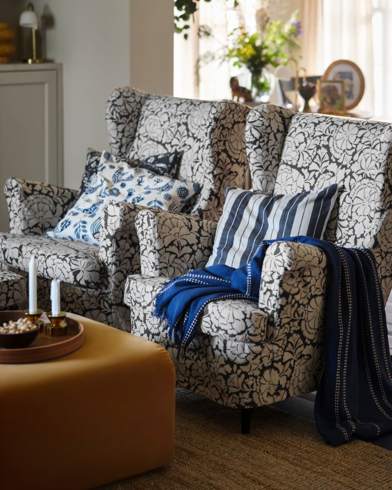 Two dark grey/beige STRANDMON slipcovers for wing chair enhance a living room, paired with patterned cushions and a throw.