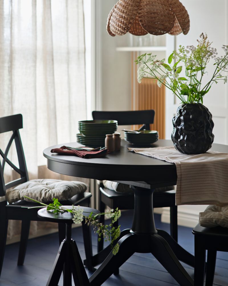 A black ROSENTORP extendable table is set with chairs, dinnerware, and a vase of flowers in a light-filled dining room.
