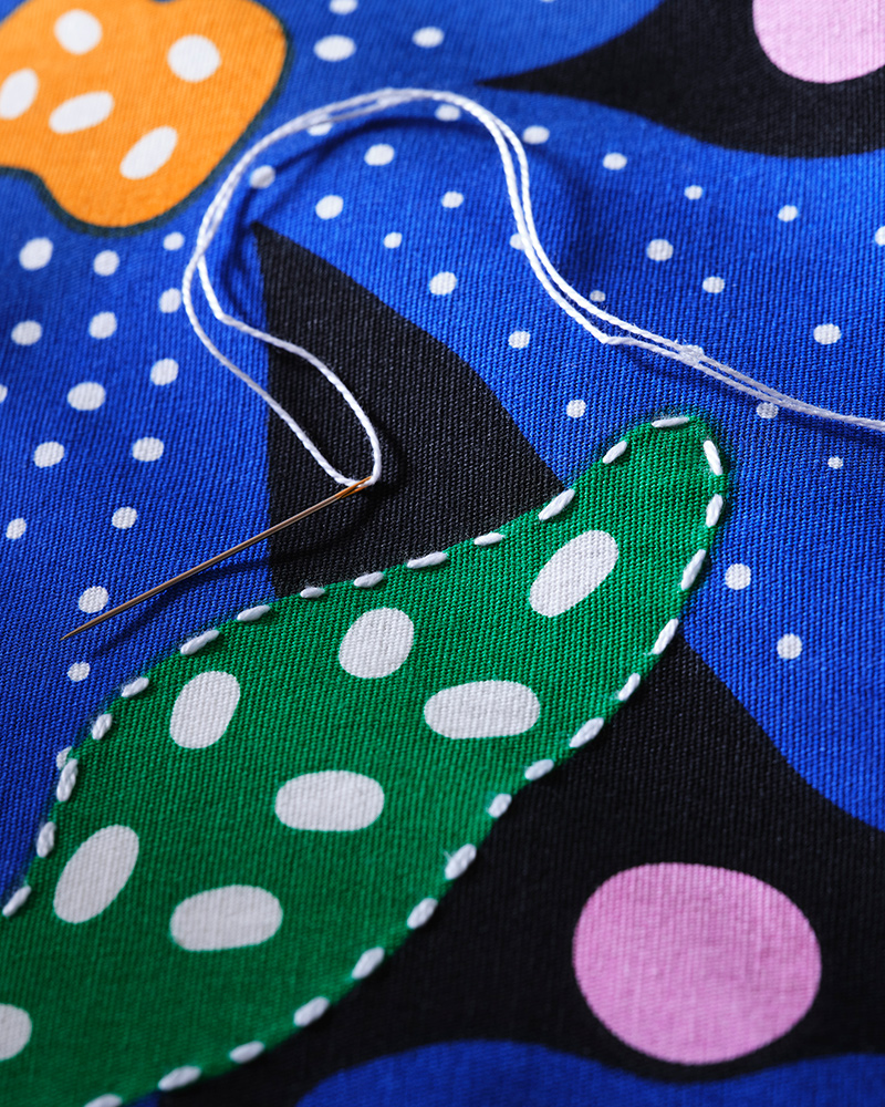 A close-up of the embroidery on the colourful floral pattern of the MÄVINN cushion cover. A needle with thread lies on top.