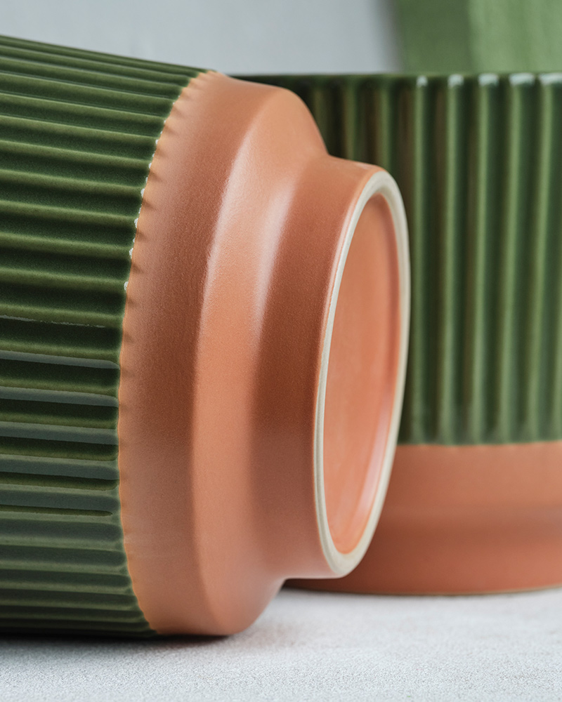 A green MÄVINN plant pot in stoneware with raised ridges and a terracotta bottom lies on its side. Behind is another pot.