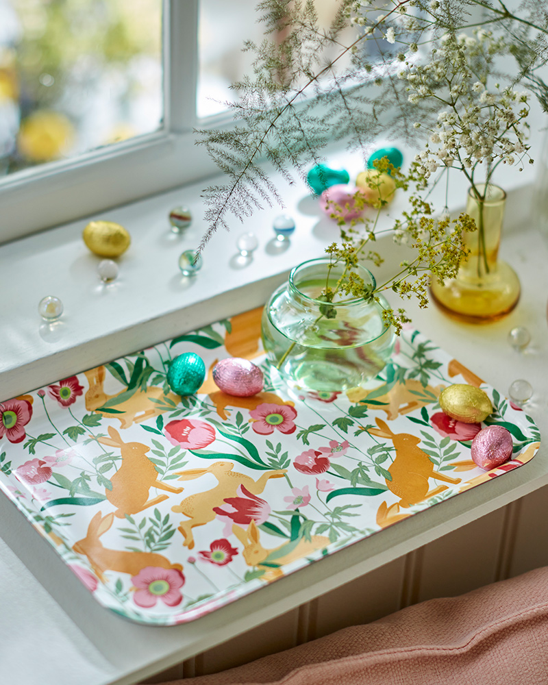 A floral-patterned TJÄRLEK tray by a window, carrying VÅRKÄNSLA chocolate eggs and a vase with SMYCKA artificial sprays.