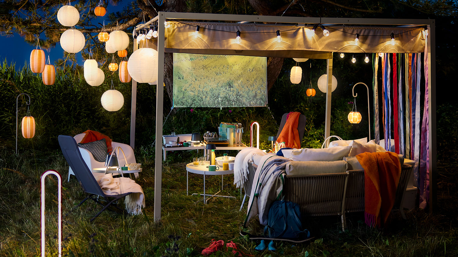 An outdoor space at night featuring striped solar-powered orange and white SOLVINDEN pendant lamps hanging from a large tree.
