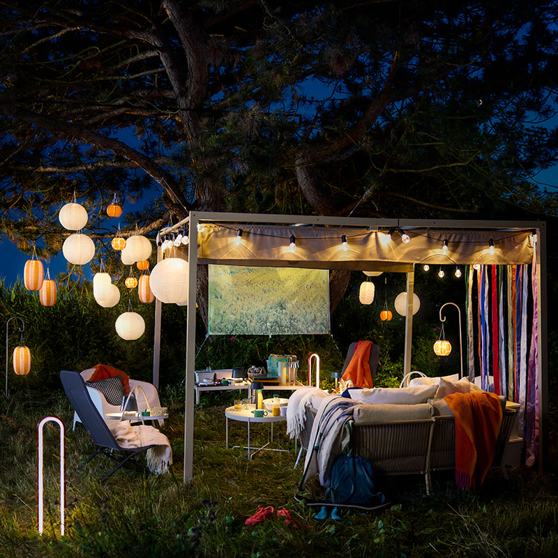 An outdoor space at night featuring striped solar-powered orange and white SOLVINDEN pendant lamps hanging from a large tree.