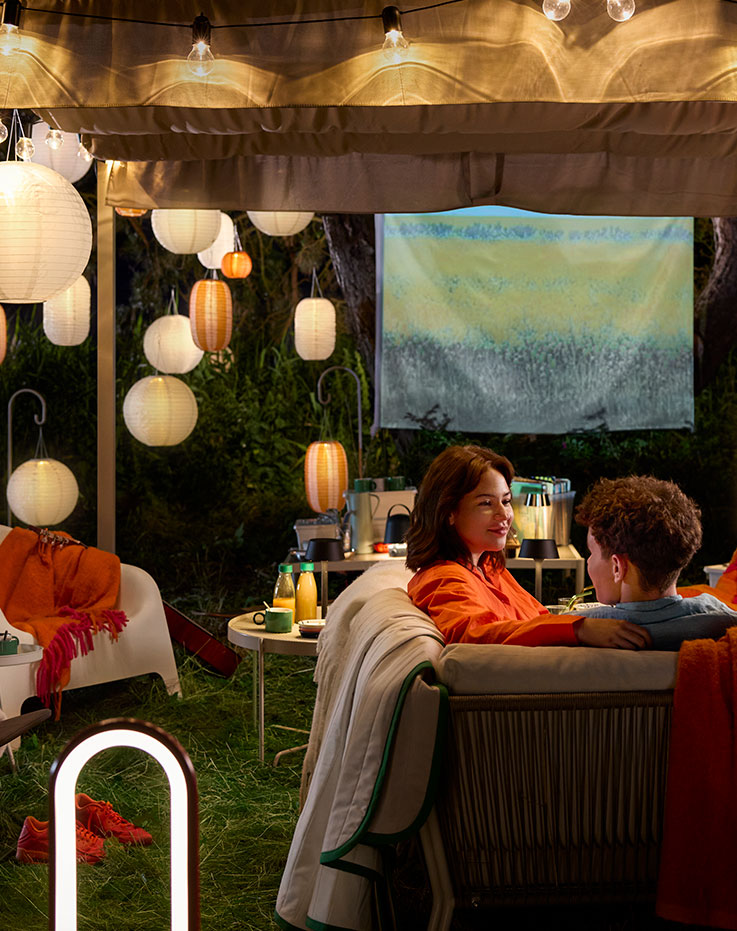 An outdoor seating area adorned with orange and white SOLVINDEN pendant lamps. Two young people in cosy blankets and cushions.
