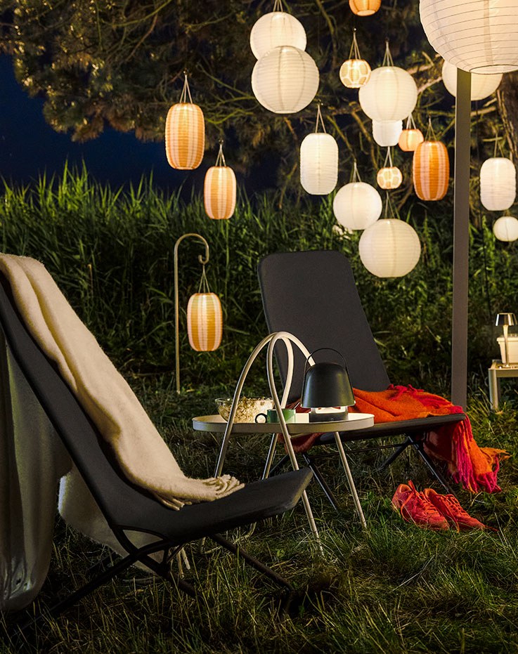 A cosy evening garden area with hanging lights features a dark grey VIGGSÖ lounge chair, surrounded by greenery.