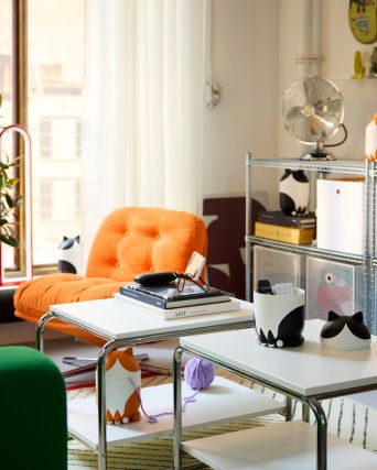 Hay tres recipientes GRESIMOJS con tapa en forma de gato sobre las mesas y estanterías de una habitación. Dos son de color negro/blanco. Uno es de color naranja/blanco.