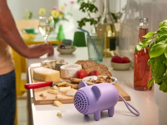 Un altavoz Bluetooth portátil GREJSIMOJS morado con forma de ratón está sobre la encimera de la cocina. Una tabla de embutidos está detrás de él.