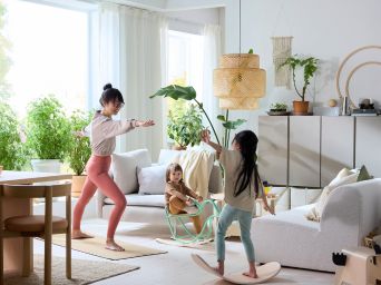 En una sala de estar, un padre practica yoga mientras sus hijos juegan en una tabla de equilibrio GREJSIMOJS y un pájaro balancín GREJSIMOJS.
