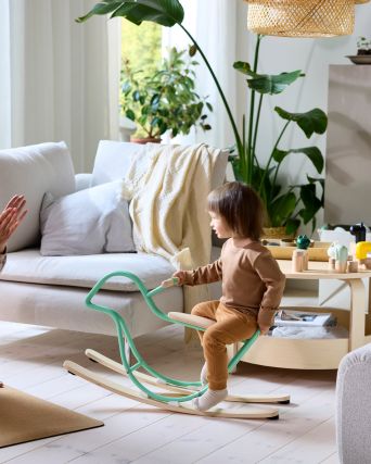 En una sala de estar luminosa y llena de plantas, un padre sobre una esterilla de yoga observa cómo su hijo se sube a un taburete GREJSIMOJS con forma de alce.