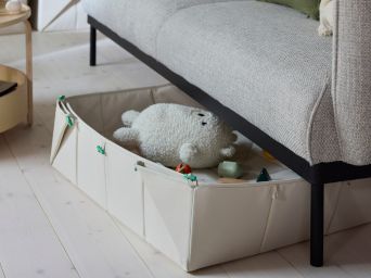 Una alfombra/caja de almacenamiento plegable GREJSIMOJS de color beige asoma por debajo de un sofá en una sala de estar moderna. En la caja hay peluches.