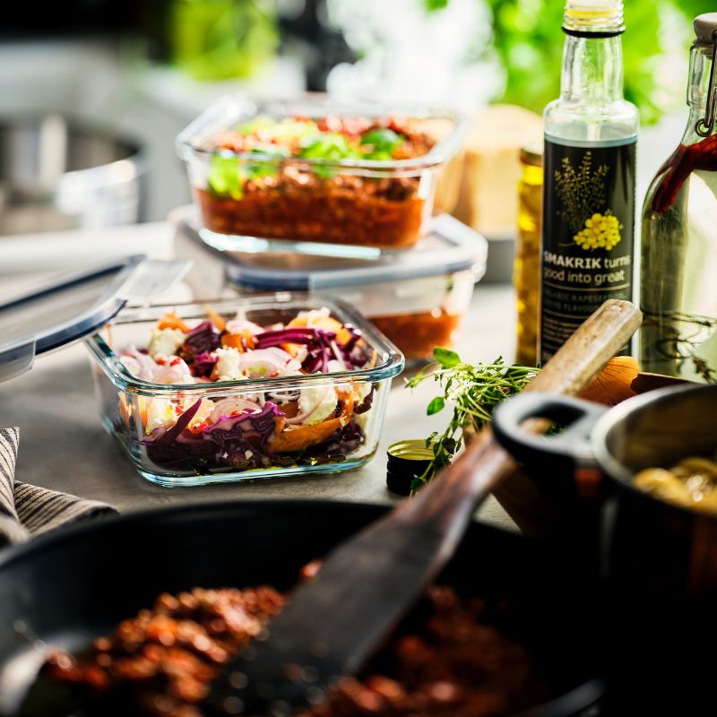A IKEA 365+ glass food storage container with prepared food is next to a pan. Two other containers are stacked behind it.