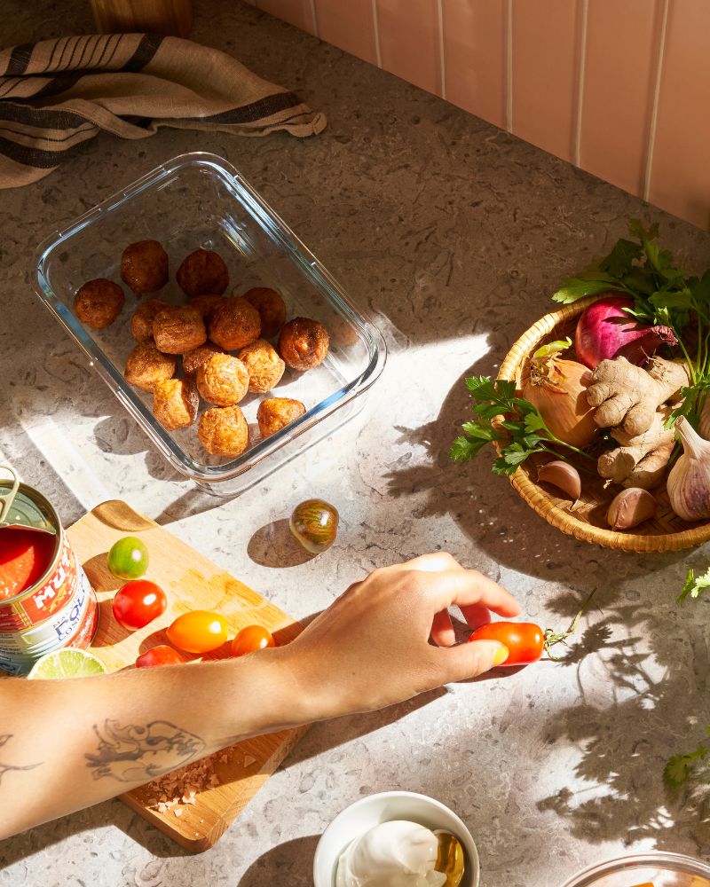 On a kitchen worktop, a person reaches for a cherry tomato. Nearby is a IKEA 365+ glass food container with plant balls.