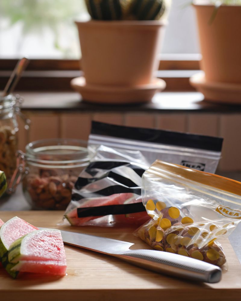 Slices of watermelon, a knife and two different sized ISTAD plastic resealable bags are on a cutting board in a kitchen.