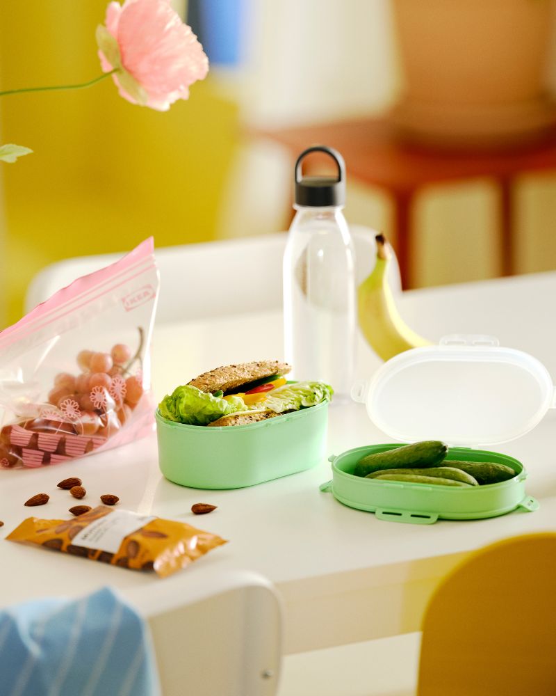 An open UTBJUDA stackable lunch box contains a sandwich in one section and pickles in the other. A water bottle is behind.