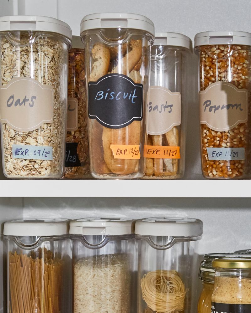 Different sized plastic food storage containers with labels populate two shelves in a kitchen cupboard.