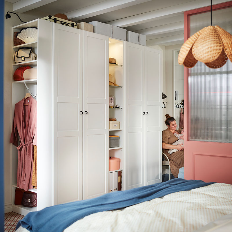 A woman and baby sit at the end of a white PAX/TYSSEDAL wardrobe with a black ÄLGANÄS clothes rack with shoe storage by it.