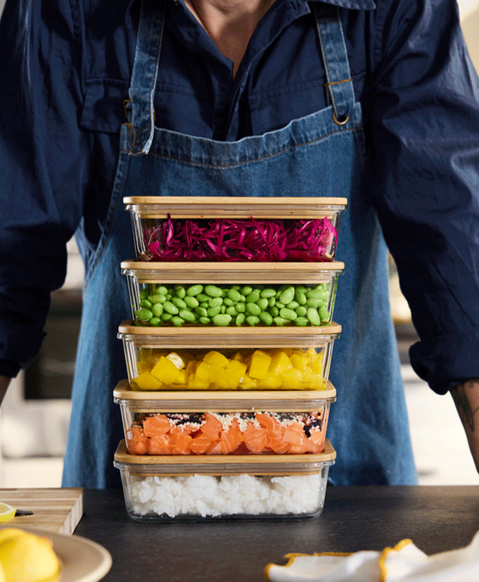 A person in a denim apron standing behind a stack of five glass food containers with bamboo lids, filled with rice, salmon, mango, and vegetables.