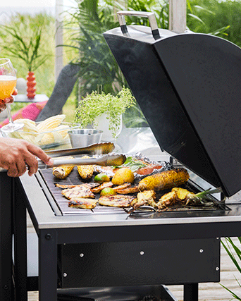 Close-up of a black charcoal barbecue grill with corn, pineapple, and food in foil cooking on the grate under an open lid.