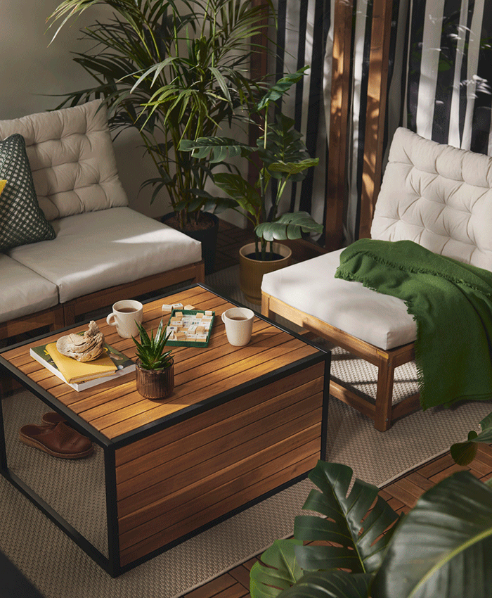 Cozy outdoor corner with wooden lounge chairs, white cushions, and a green throw, centered around a wooden coffee table with tea and games.