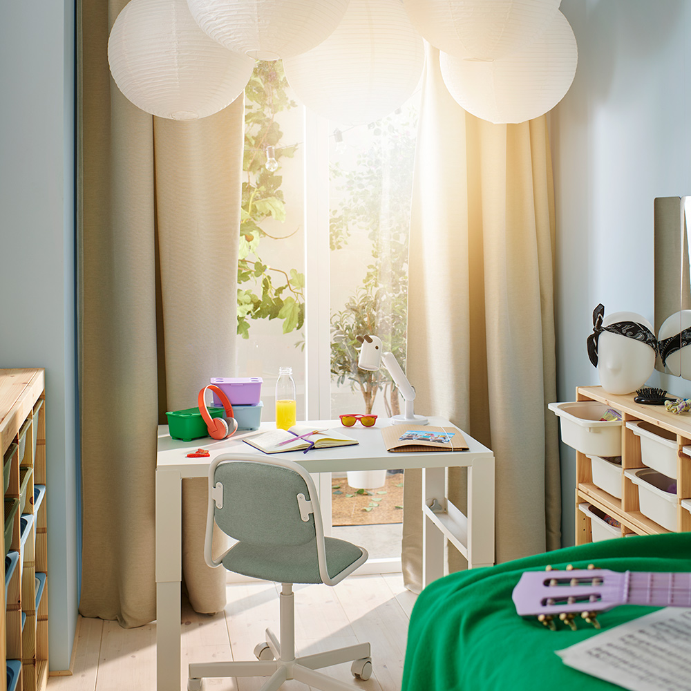 Un escritorio PÅHL blanco en una habitación luminosa y soleada que tiene cajas GLIS verdes, lila y azules, y una lámpara de trabajo LED KRUX blanca encima.