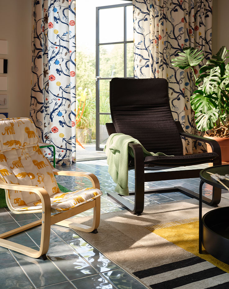 A black-brown POÄNG armchair next to an animal patterned smaller POÄNG armchair in a room with plants and patterned curtains.