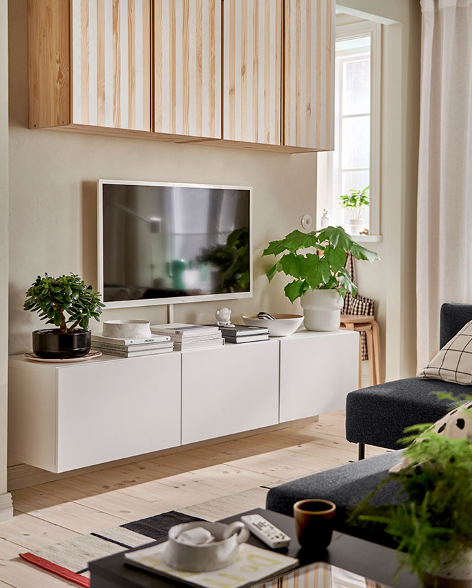 A white BESTÅ TV bench with doors is mounted on a wall underneath a wall-mounted TV and IVAR cabinets painted with stripes.