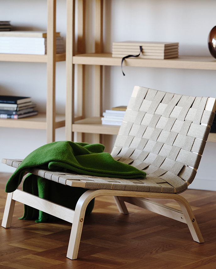A living room with a green STOCKHOLM 2025 easy chair made of birch veneer with a woven seat, accompanied by a folded throw.