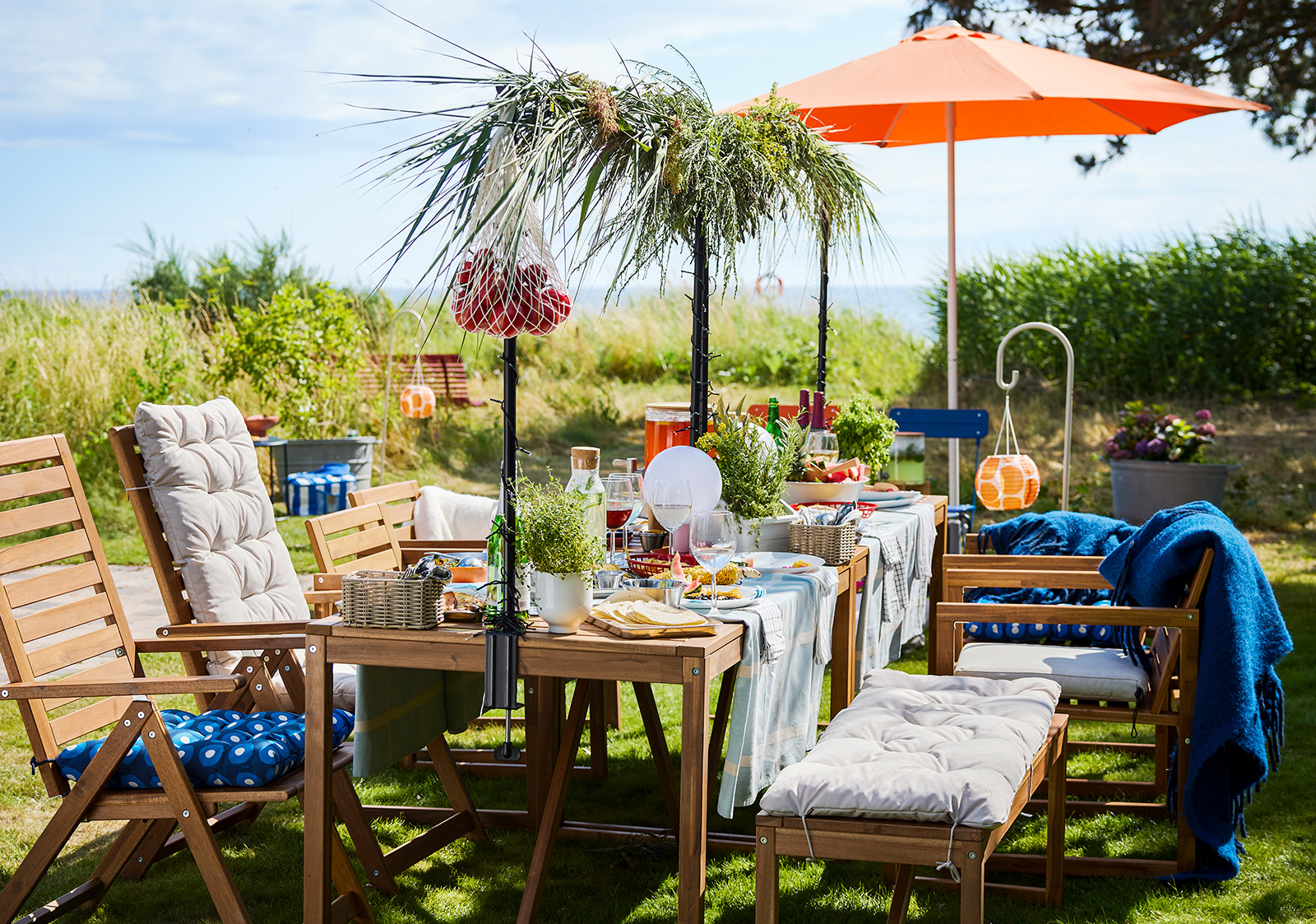 A garden setting with a light brown wooden NÄMMARÖ table with food and drinks on a sunny day. Chairs, a bench and a parasol.