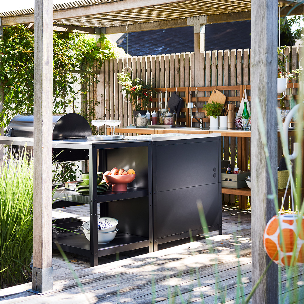 A large wooden patio with plants and lamps and a black GRILLSKÄR charcoal barbecue, part of a kitchen island setup.