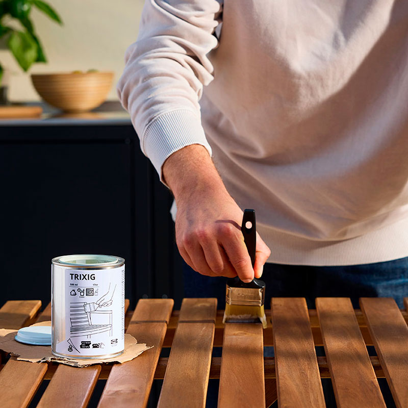 A person is re-staining a wooden outdoor table with a brush next to a can of TRIXIG outdoor wood stain.